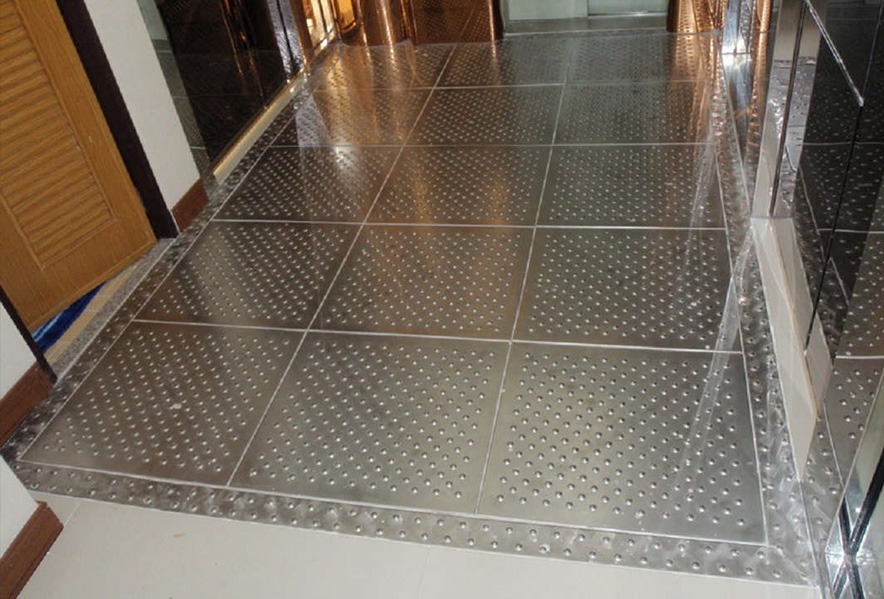 A hallway floor with large metallic tiles, some featuring raised circular patterns and others smooth, reflecting light from surrounding surfaces.