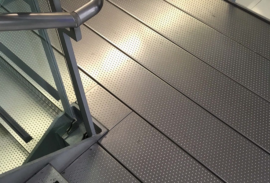 A close-up view of a metal staircase and floor with a textured, dotted surface for slip resistance; horizontal lines and metallic railings are visible.