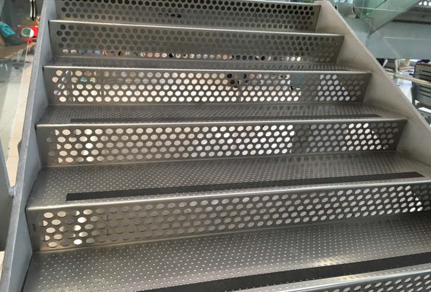 Close-up view of modern metal stairs with perforated, circular holes on each step, giving the staircase a distinctive, industrial look.