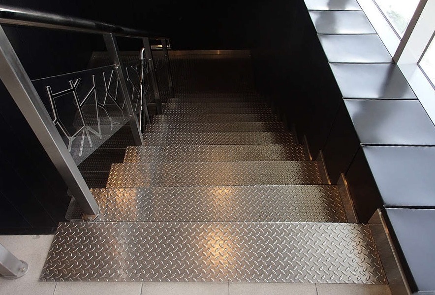 A staircase with metallic, diamond-plate patterned steps and a metal railing, descending between dark walls. There are window-side bench seats with black cushions to the right.