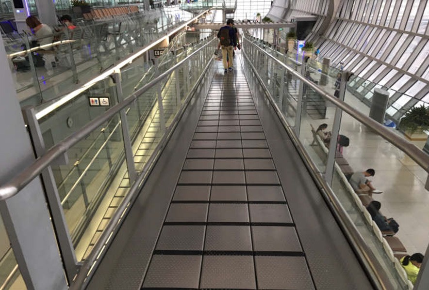 A walkway with metal railings in an airport terminal, leading toward a person with luggage. People are sitting and walking in the area below to the right, and large windows line the curved wall.