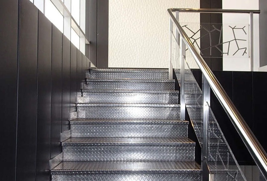A metal staircase with diamond plate steps, black side panels, and a shiny metal handrail ascends toward a bright window and a textured white wall.