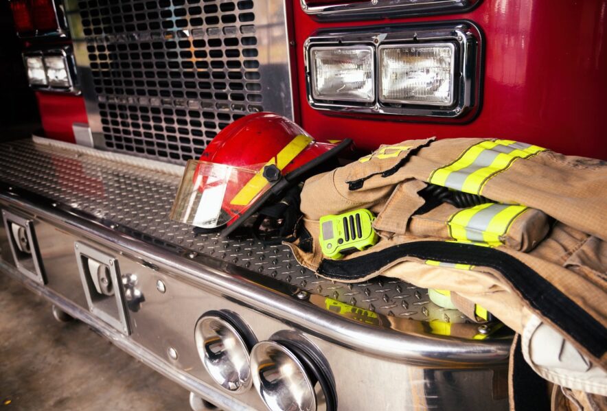A firefighter’s helmet, uniform, and a radio are placed on the metal bumper of a red fire truck, with headlights and part of the front grille visible in the background.