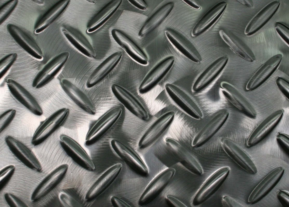Close-up of a metallic diamond plate with a raised, repeating pattern of oval shapes, giving the surface a textured, industrial appearance. The metal reflects light, creating shiny and matte areas.
