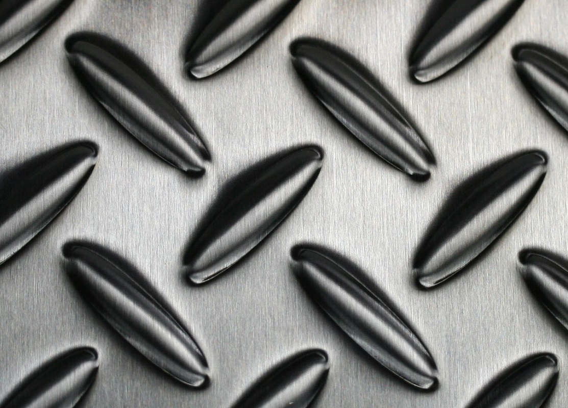 Close-up of a metallic surface with a raised diamond plate pattern, featuring diagonal, oval-shaped ridges in a repeating design on a brushed steel background.