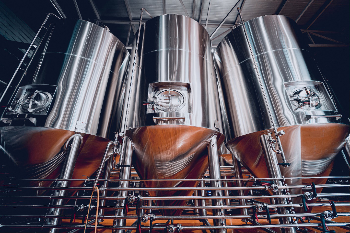Three large, shiny stainless steel fermentation tanks stand side by side in a brewery, with pipes and valves connected at the bottom, reflecting the industrial environment.