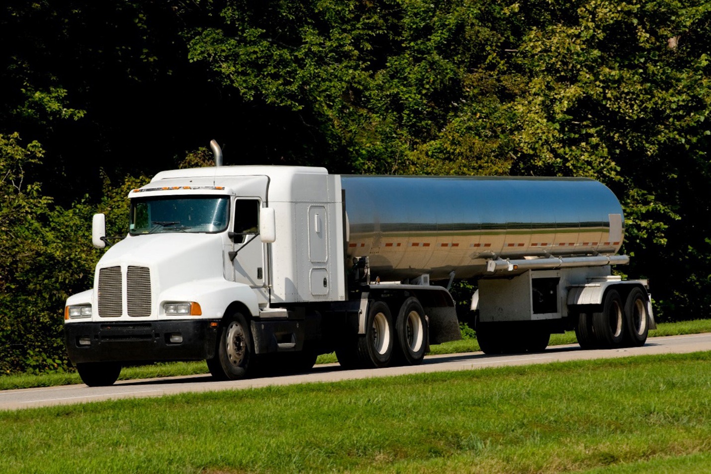A white semi-truck with a large silver tanker trailer drives down a road bordered by green grass and dense trees in the background.