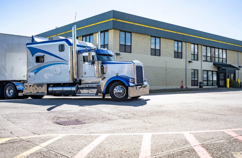 A shiny blue and white semi-truck parked outside a modern industrial building with large windows and a yellow trim. The scene is bright and sunny.