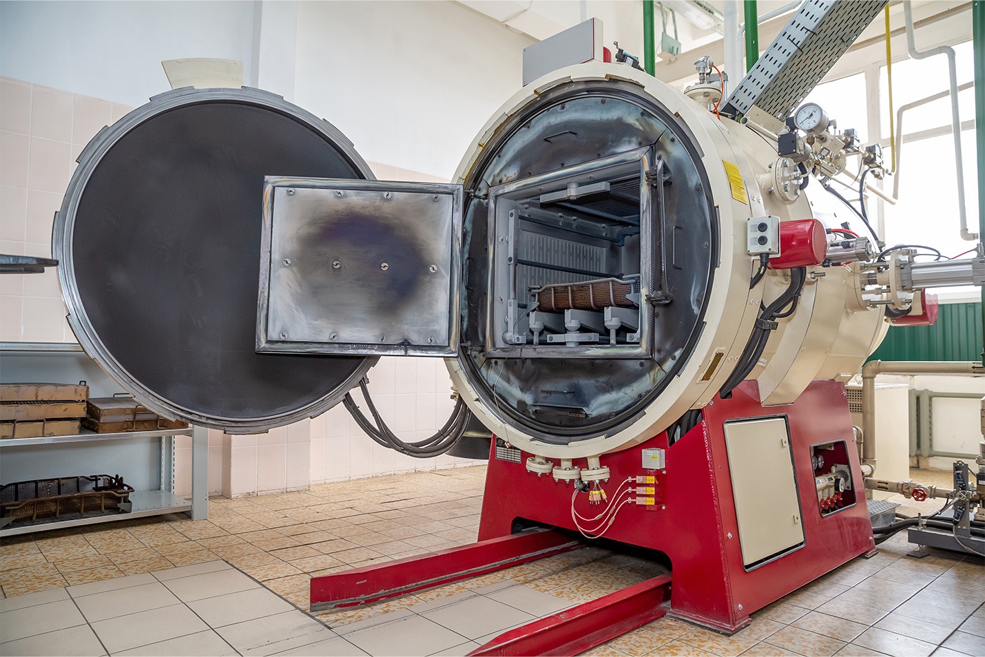 A large industrial vacuum furnace with its round front door open, revealing a metal chamber inside. The machine is red and white, situated in a clean, tiled laboratory or workshop environment.